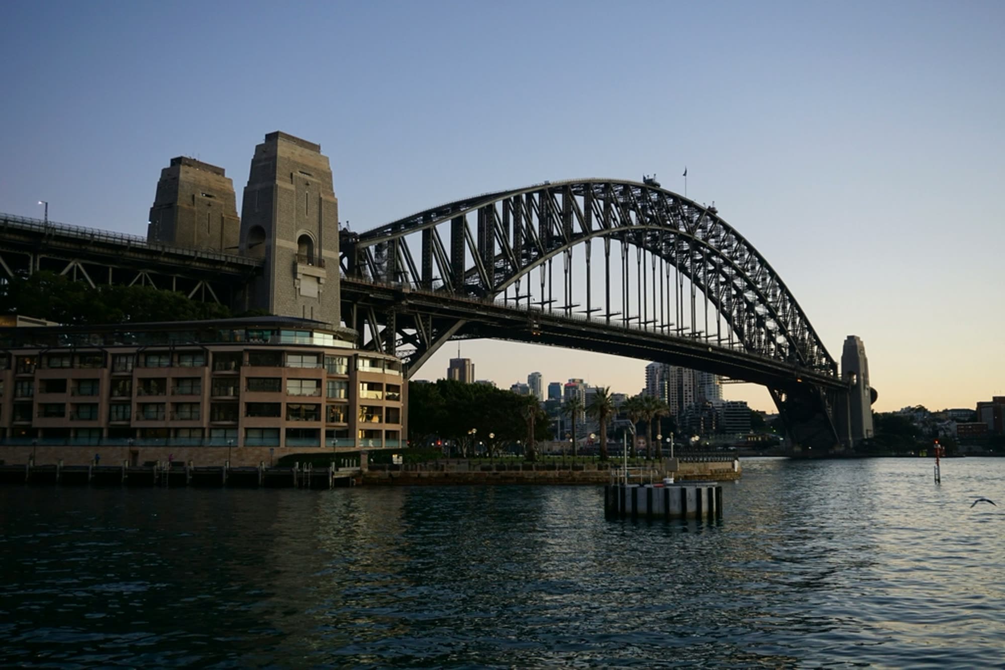 A steel arch harbour bridge at dusk seen from a quay
