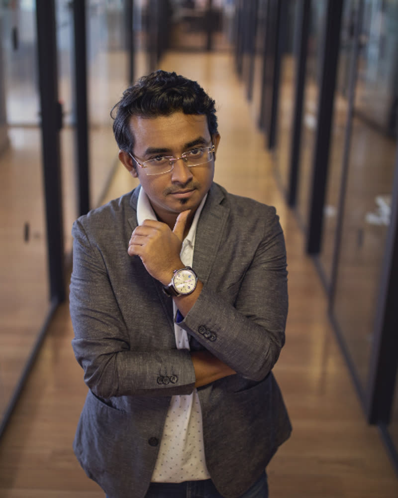 Portrait of Rohan Mehta, a man with dark hair and glasses in a grey jacket standing in a glass-walled corridor
